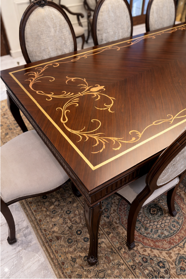 Wooden dining table with intricate gold inlay design, surrounded by chairs on a patterned rug.